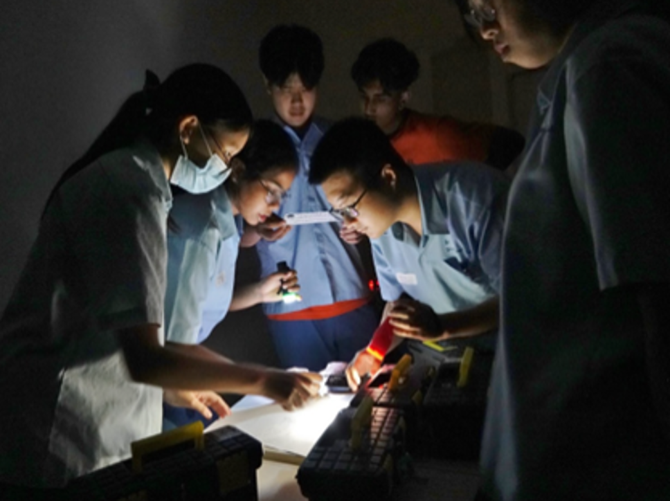 Students gather in a dimly lit room using flashlights to examine objects closely during a hands-on investigative or science activity.